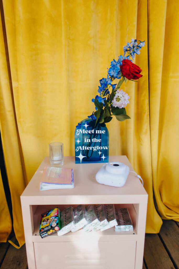 A table at a wedding venue with a sign reading 'Meet me in the Afterglow,' a bouquet of flowers, and a white instant camera, against a vibrant yellow curtain.