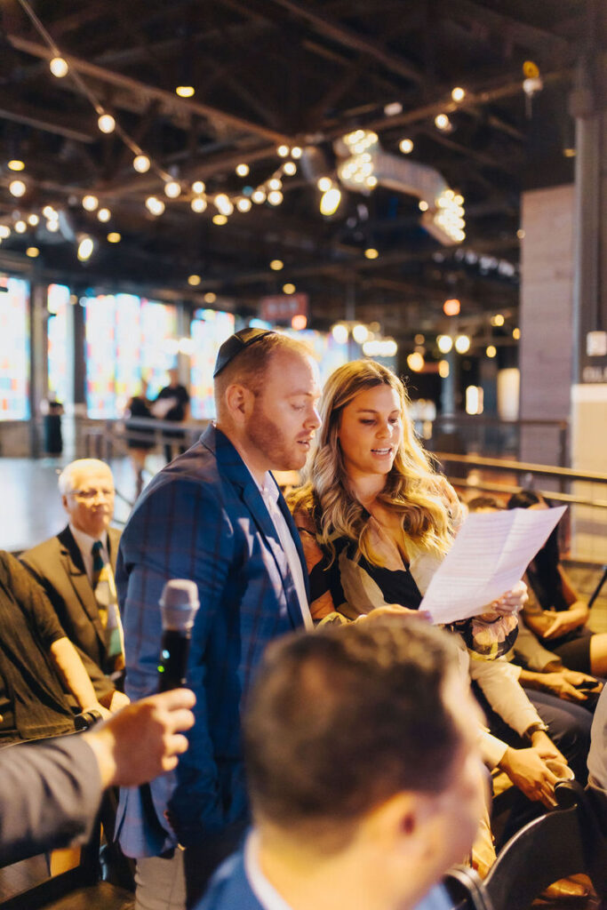 A couple standing and reading from a sheet of paper at a wedding event, with guests and string lights visible in the soft-focus background of an urban loft space.