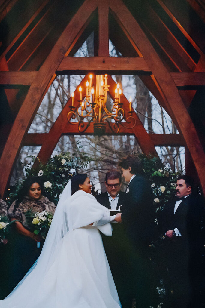 An intimate wedding ceremony with a couple at an altar under an ornate chandelier, framed by a wooden arch and surrounded by guests