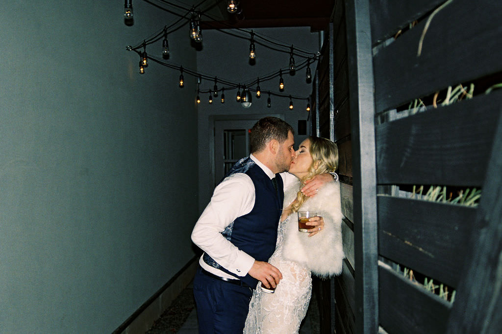 Newlywed couple share a private, joyful kiss in a dimly lit corridor adorned with string lights, creating an intimate post-ceremony moment.