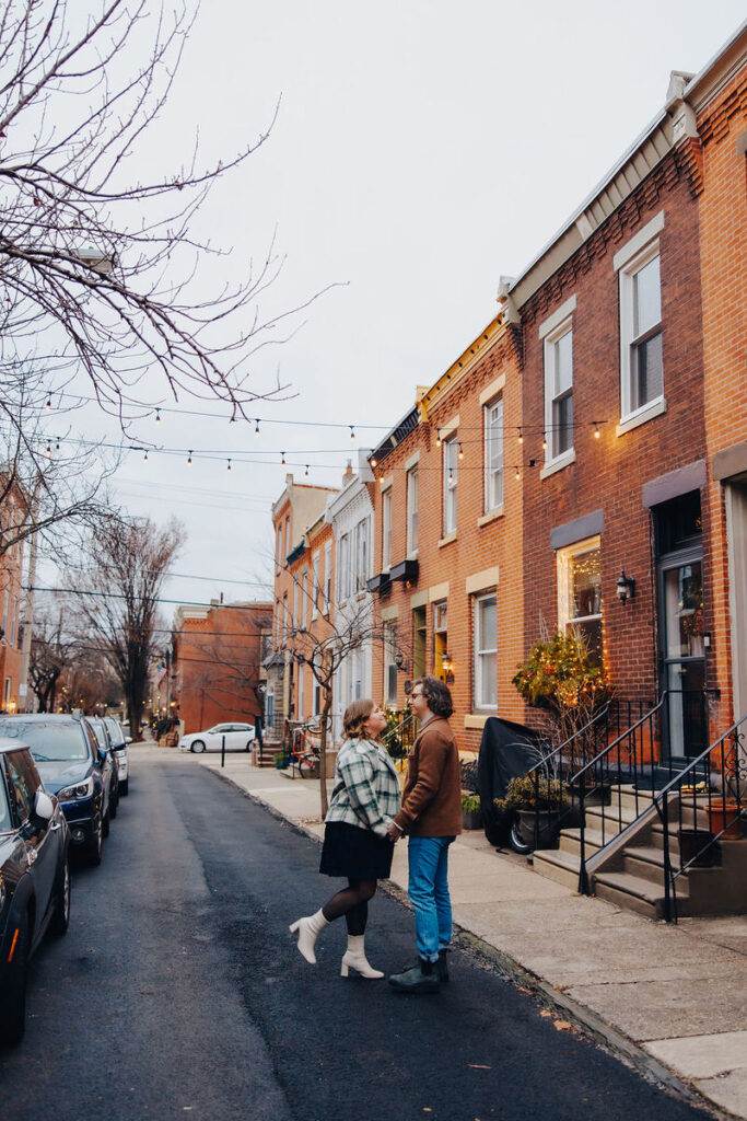 A couple holding hands and walking down a city street lined with brick buildings and festive string lights