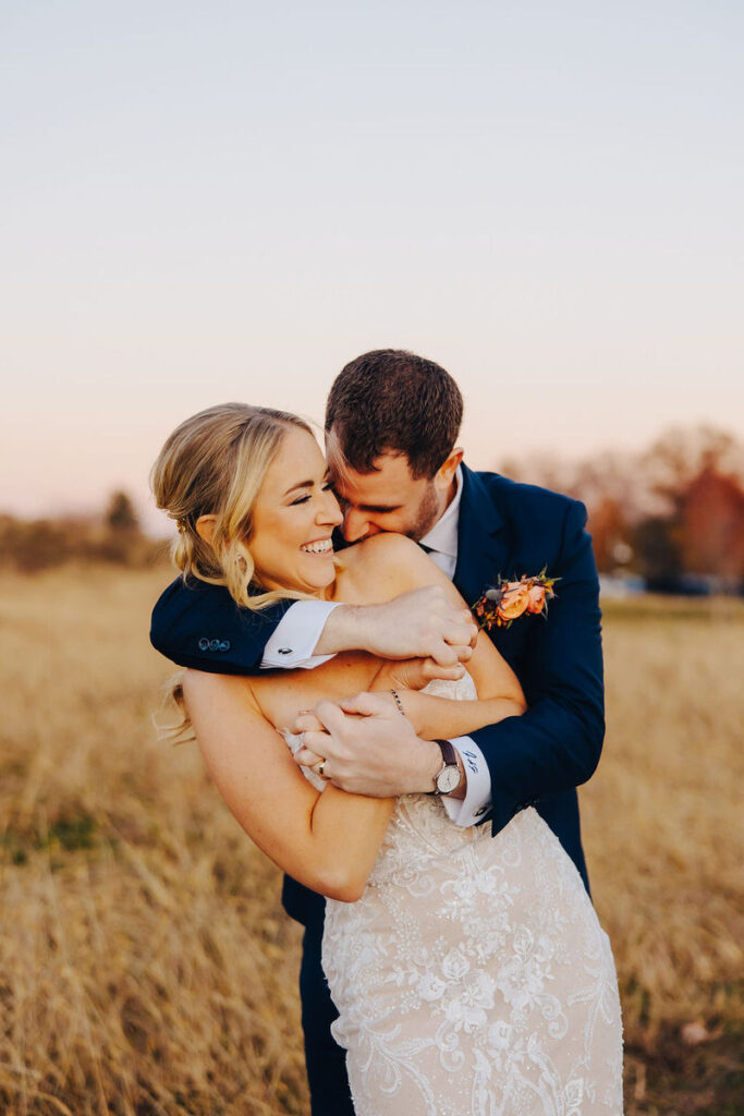 Digital photo of a smiling couple, the one hugging the other from behind, amidst a golden field at dusk, expressing warmth and joy