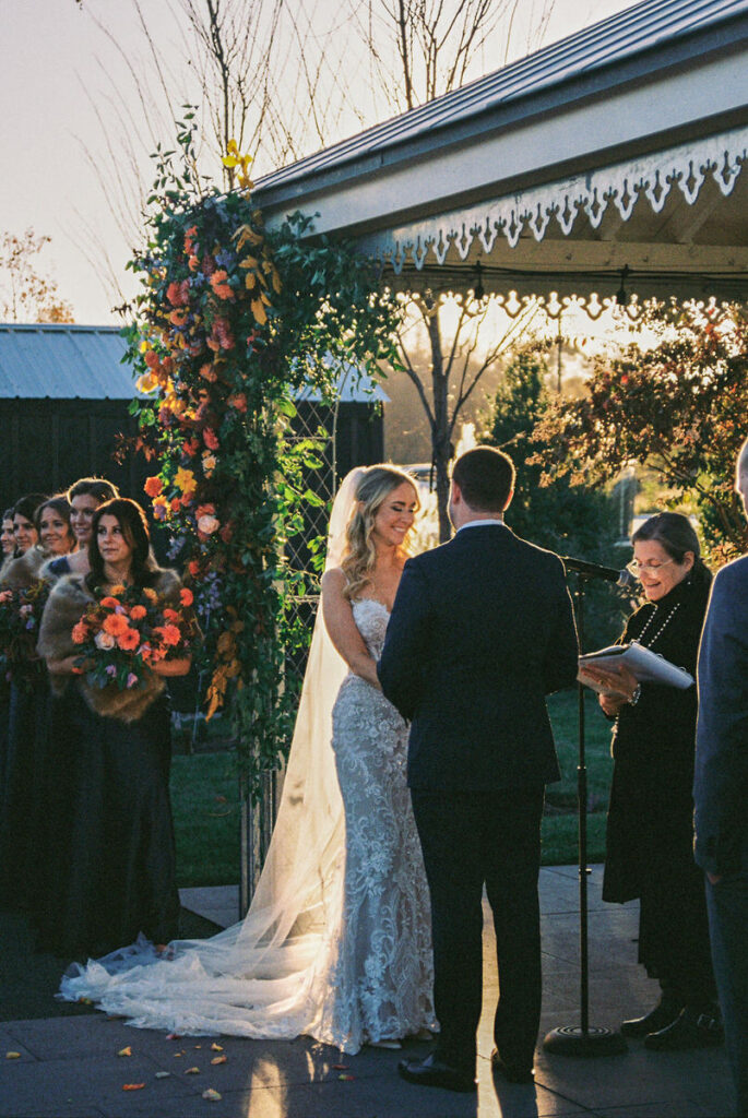 A tender moment captured on film, where a couple hold hands under a gazebo with guests looking on during an outdoor ceremony