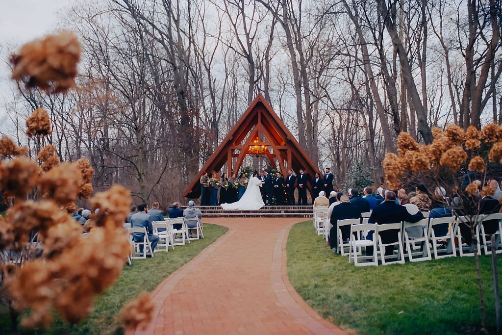A grand outdoor wedding scene with guests seated facing a striking wooden chapel-like structure, the couple exchanging vows under a triangular arch
