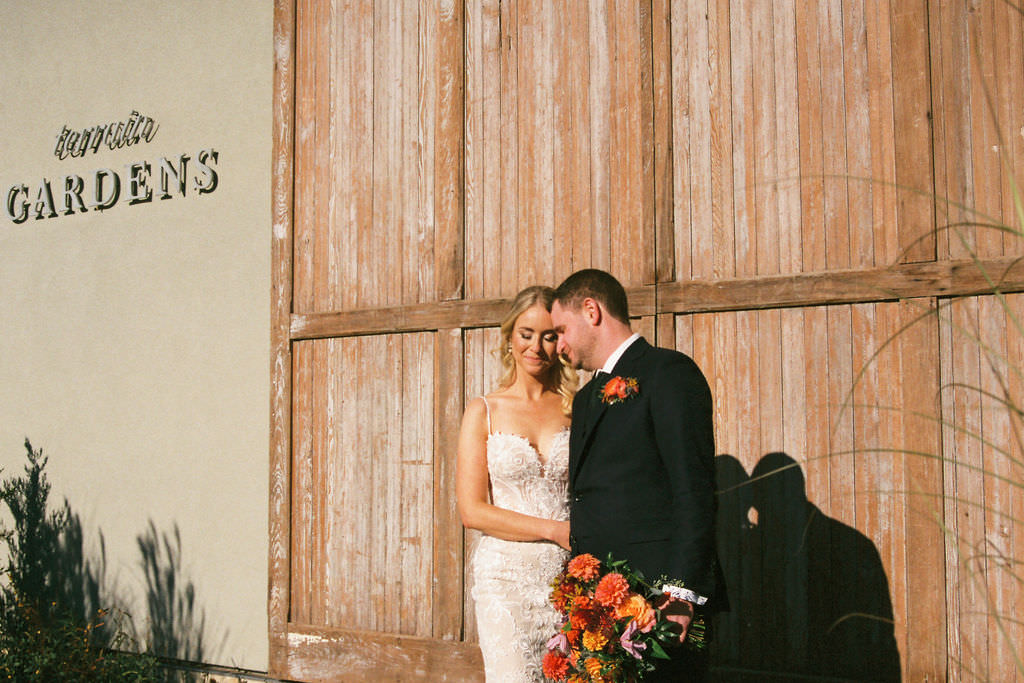 Film wedding photography showing a newlywed couple standing lovingly together in front of a rustic wooden backdrop with 'beautiful gardens' text on it.