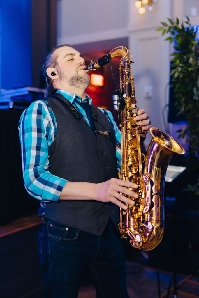 A male saxophonist in a blue checkered shirt playing passionately at an event, with a soft focus on the brass instrument.