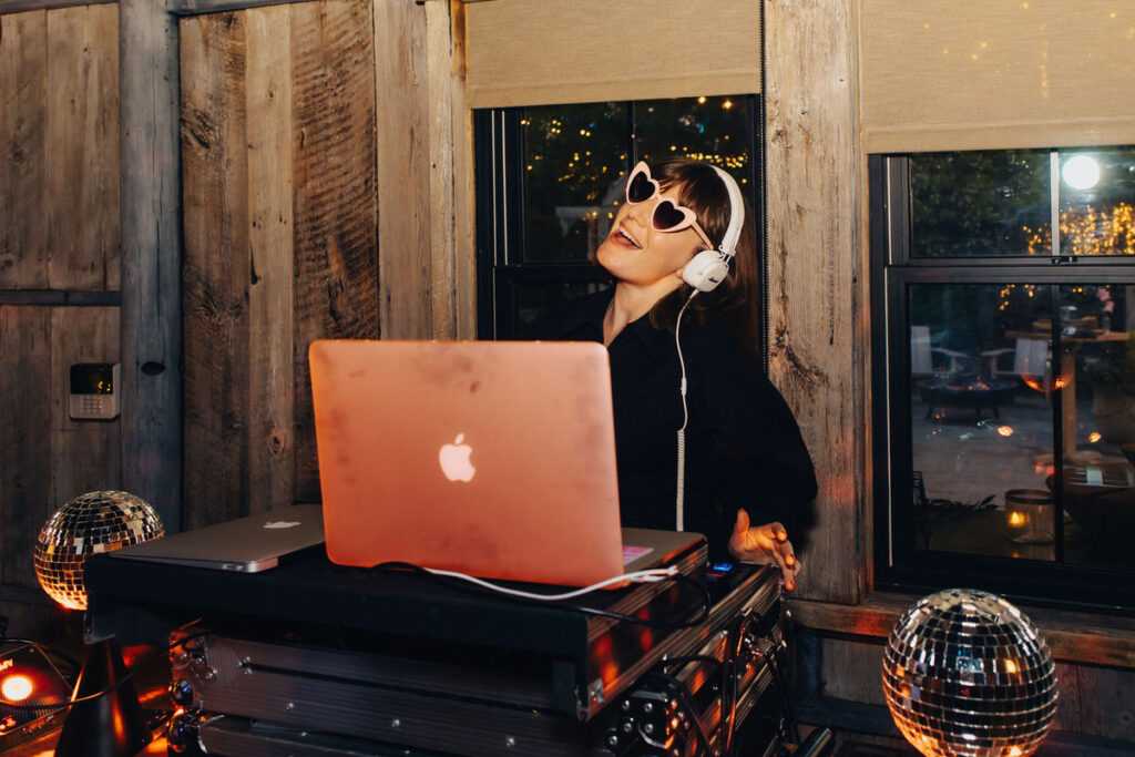 A female DJ wearing heart-shaped sunglasses and headphones, selecting tunes on her laptop at a festive event with a disco ball in the foreground.