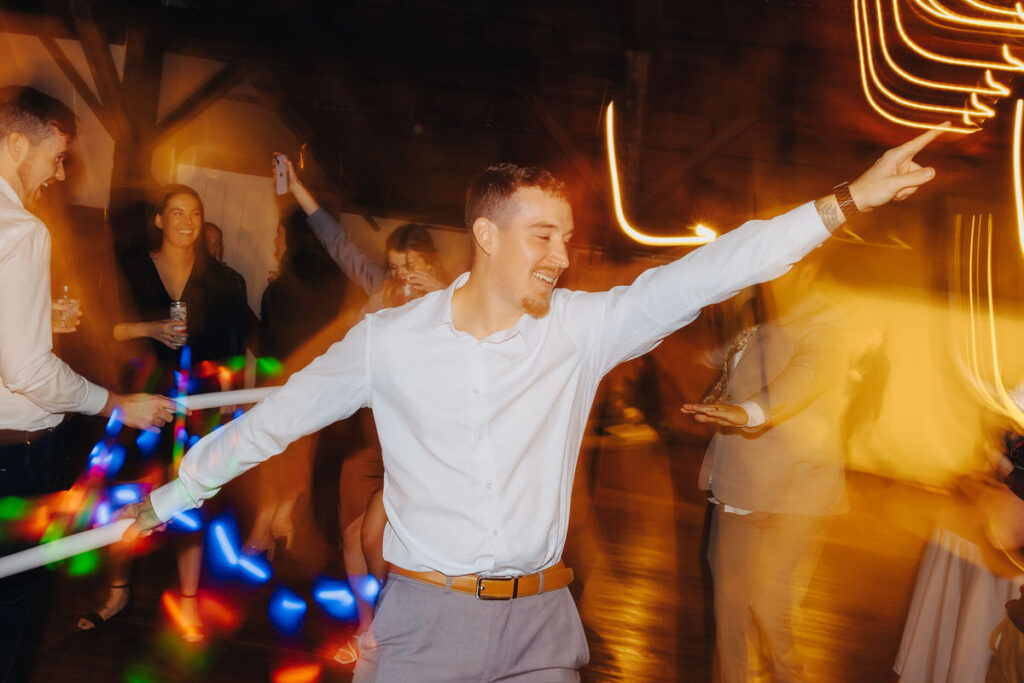 A man in a white shirt joyfully dancing with arms outstretched at a vibrant party, surrounded by colorful light trails