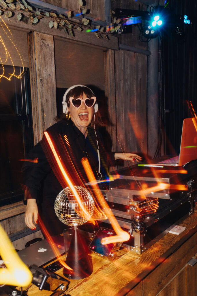 A female DJ wearing heart-shaped sunglasses and headphones playing music at an event, with a disco ball and colorful light effects.