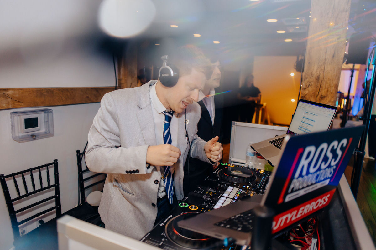 A male DJ in a grey suit and blue tie getting into the groove while mixing tracks at a wedding party.