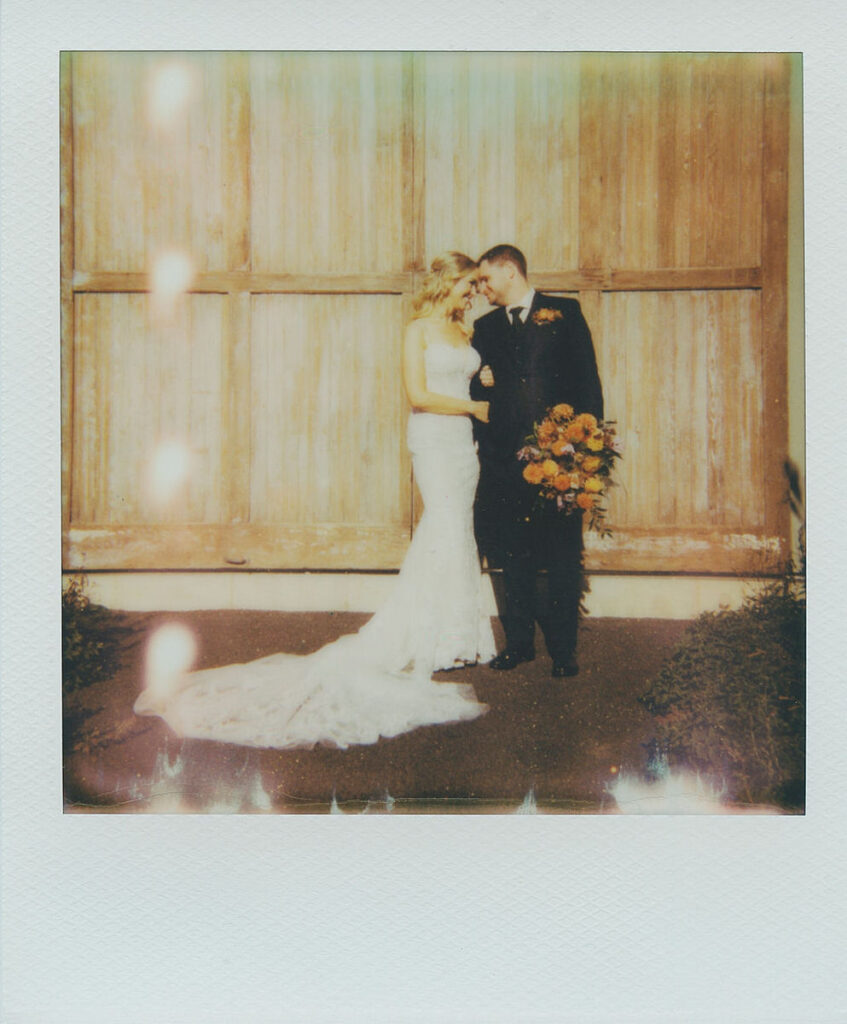 A newlywed couple embracing while standing in front of rustic wooden doors, with the bride holding a bouquet of orange flowers, captured on a Polaroid with light flares