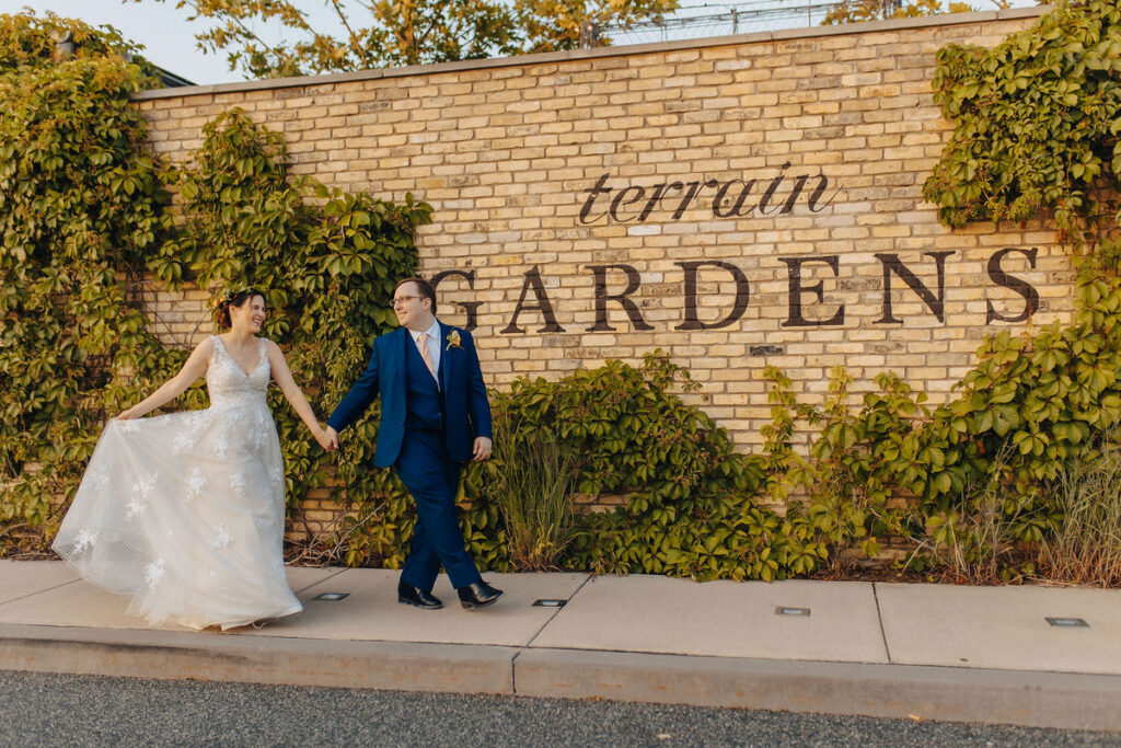 A joyful bride and groom holding hands and walking past a brick wall with "terrain gardens" signage, surrounded by ivy, in a casual yet elegant wedding setting.