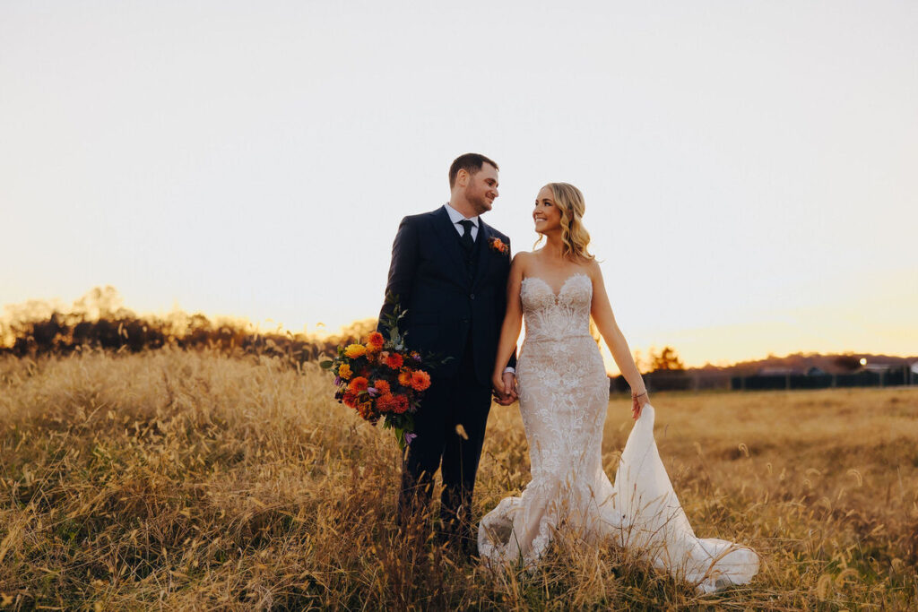 A bride and groom walking hand-in-hand through golden fields at sunset, the warm glow highlighting their affectionate smiles and the bride's vibrant bouquet.