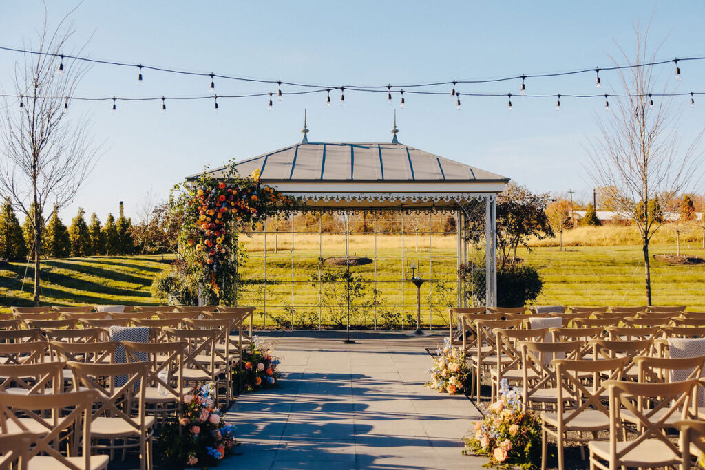 An outdoor wedding ceremony setup featuring a gazebo adorned with vibrant flowers, string lights above, and rows of beige chairs against a backdrop of open skies and greenery.