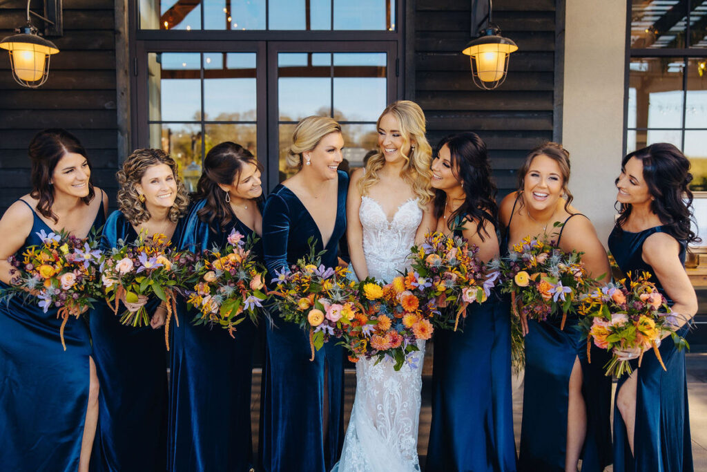 A radiant bridal party in navy blue dresses, holding colorful bouquets, gathered around the bride in a lace dress, sharing a moment of laughter and excitement.