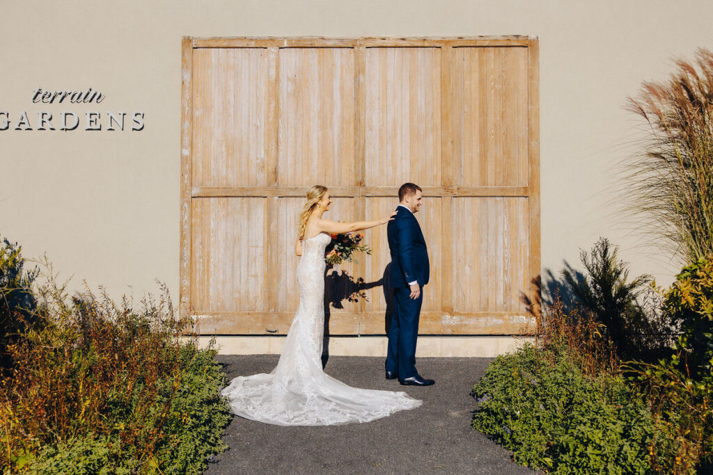 A newlywed couple playfully interacting in front of a large wooden barn door with "terrain gardens" signage, under a clear blue sky, encapsulating a rustic wedding charm.