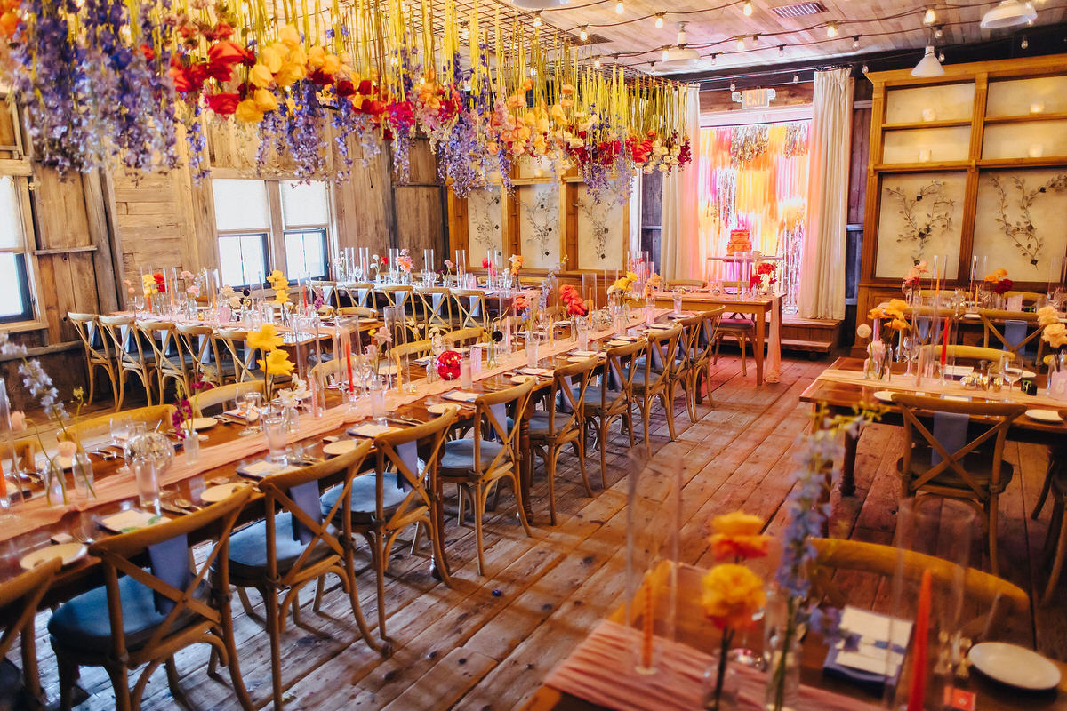 An indoor reception area lavishly decorated with hanging floral arrangements in a rustic barn setting, wooden tables set for a celebratory feast, embodying a bohemian elegance.