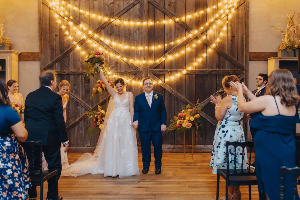 The bride leading a joyful procession out of a rustic barn venue, with guests clapping and string lights creating a starry effect, symbolizing the cheerful beginning of married life.