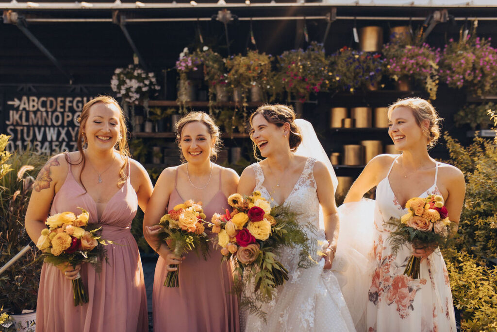 A group of bridesmaids in flowing pink dresses, laughing and posing with the bride, who is adorned in a white lace dress, against a backdrop of vibrant flowers and rustic decor.