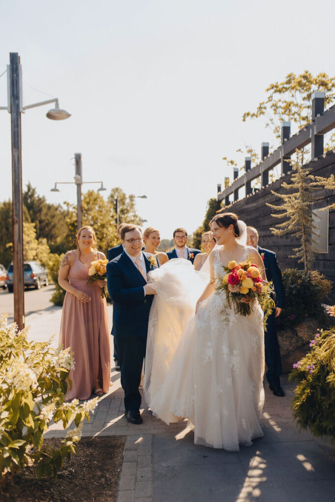 A bride in a flowing white gown and groom in a navy suit walk down the sidewalk, smiling, flanked by bridesmaids in pink dresses and groomsmen in suits, with urban street elements in the background.