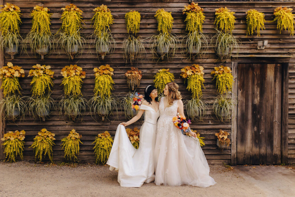 Two brides in elegant white wedding gowns stand before a rustic wooden wall adorned with cascading yellow flowers, sharing a moment of laughter and connection.