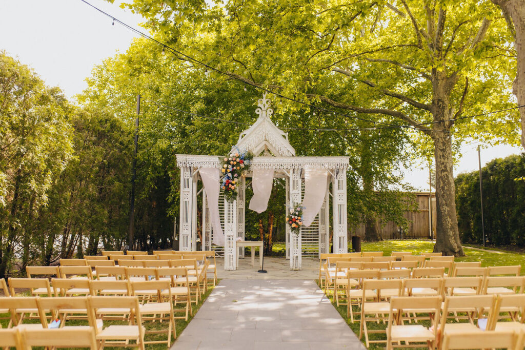 An outdoor wedding venue with a white gazebo decorated with flowers and draped fabric, wooden chairs lined up for guests, set under a canopy of green trees.