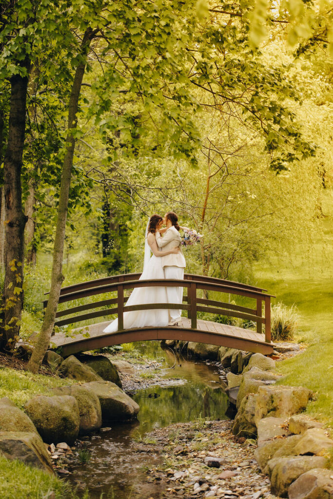 A bride kissing her partner's forehead as they stand on a small bridge over a stream. 