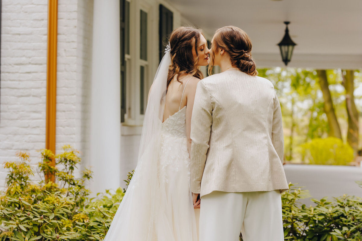 Two brides kissing with one in a flowing white gown and the other in a white pantsuit, set against a white house with greenery at their Historic Ashland wedding