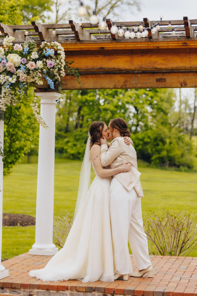 Two brides kissing under a pergola during their wedding ceremony. 