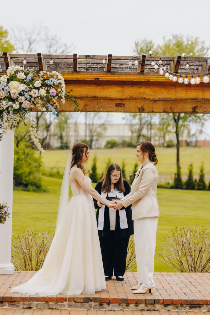 Two brides holding hands as they say their vows during their wedding ceremony. 