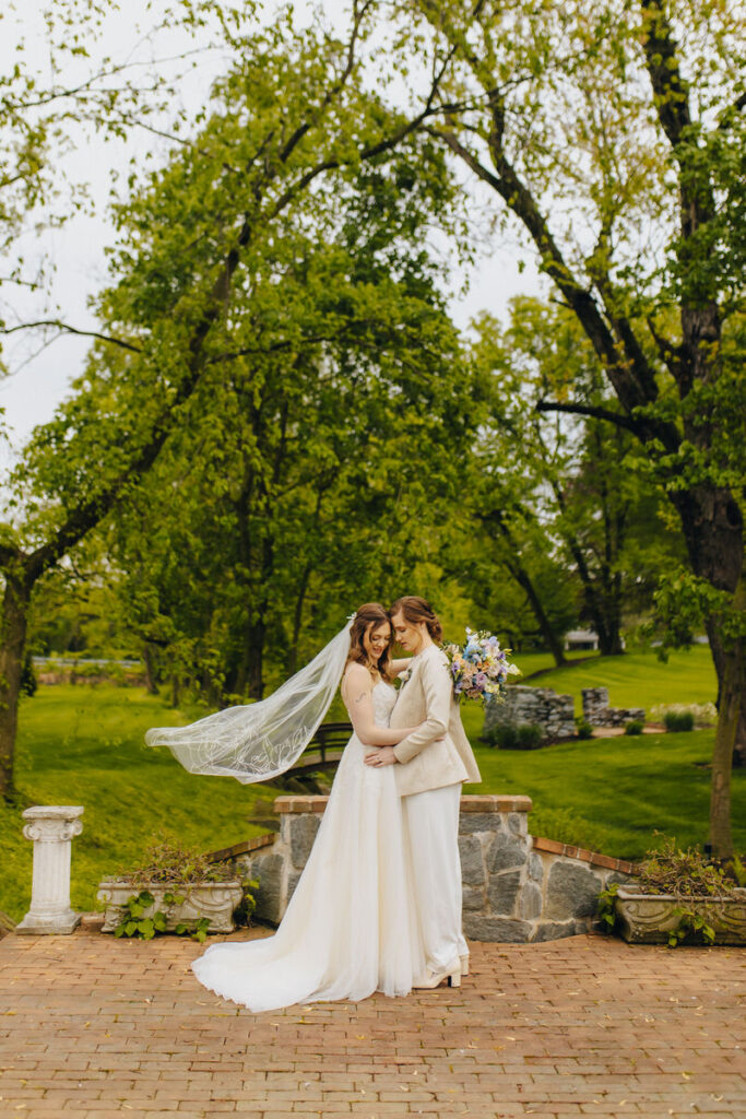 Two brides hugging closely in a garden area as one of the veils flows in the wind. 