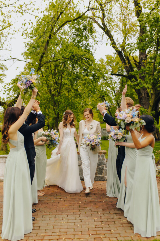 Two brides walking through a tunnel of their wedding parties with their hands up. 