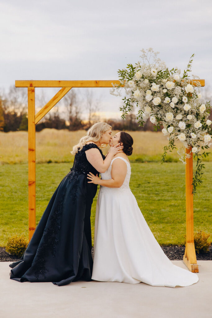 Two brides during their first kiss with a flower covered wooden arch behind them. 