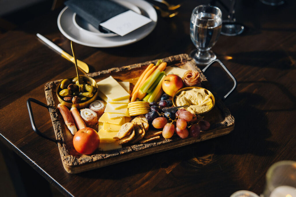 A rustic charcuterie board filled with an assortment of cheeses, meats, fruits, and pickles, arranged on a wooden table at a wedding reception