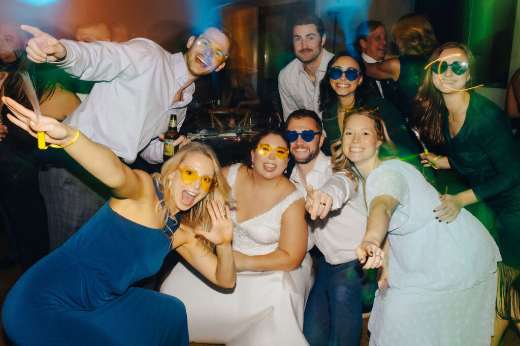 A lively group of wedding attendees wearing fun, colorful sunglasses and dancing