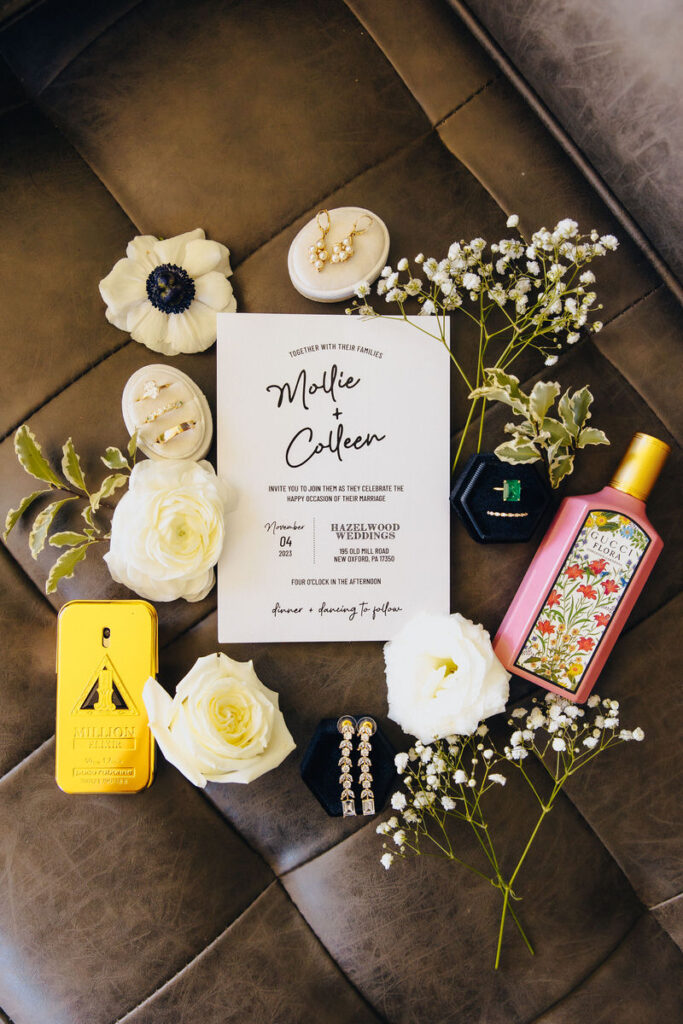 A neatly arranged wedding invitation surrounded by wedding day accessories including white flowers, a gold perfume bottle, and jewelry, on a dark leather background