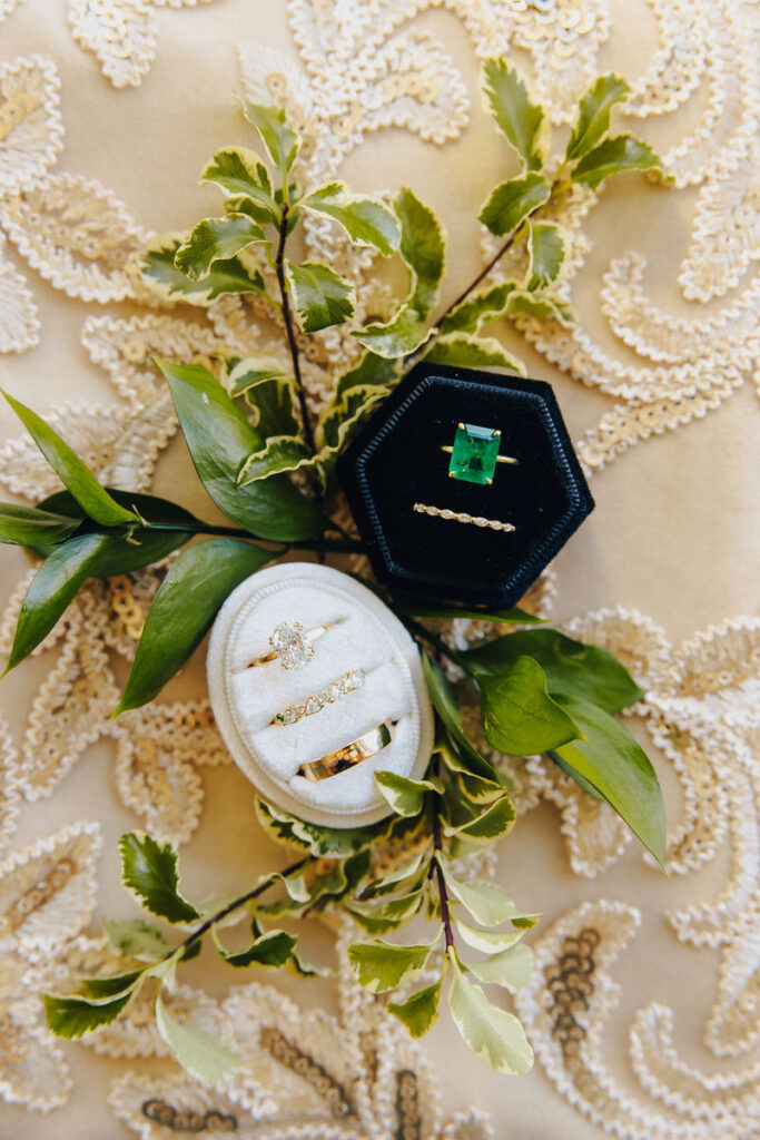 A close-up of wedding rings and a vibrant green emerald engagement ring presented in velvet boxes among fresh green leaves and ivory lace fabric