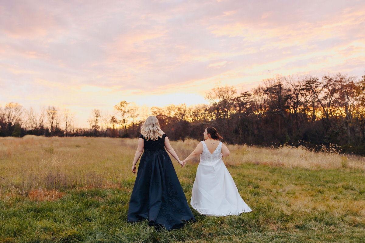 Two brides seen from the back, walking hand-in-hand into the sunset across a scenic field in the middle of their perfect wedding timeline with a first look.