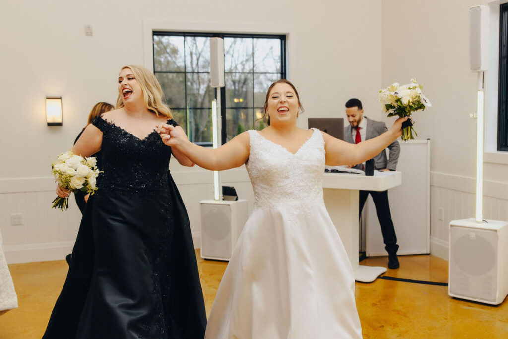 Two brides joyfully dancing together at their wedding reception, with one in a white gown and the other in a black dress