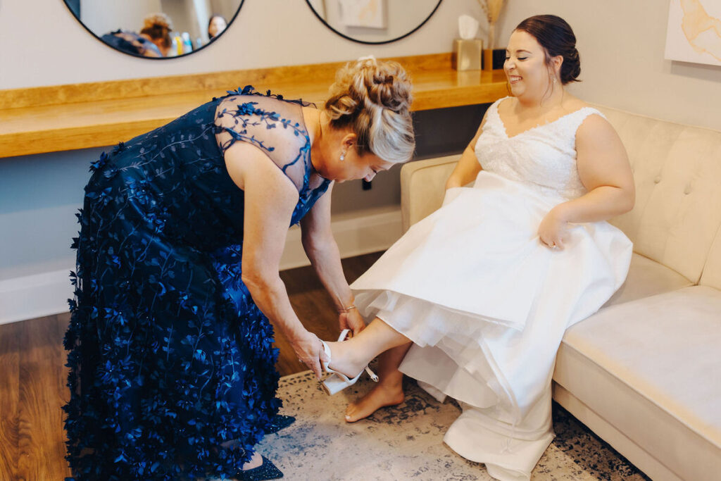 A mother assists the bride with her shoes, the bride is smiling and seated on a cream sofa, with the mother's blue dress detailed with floral appliqué