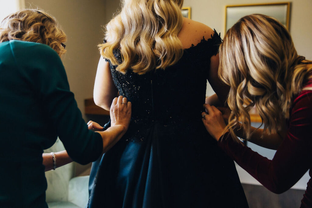 A behind-the-scenes moment as two women, one in a green dress and another in a red dress, assist a bride in a black dress with getting dressed