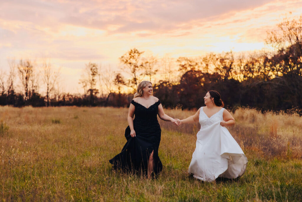 Two brides holding hands and walking through a field during golden hour, with a beautiful sunset providing a warm backdrop