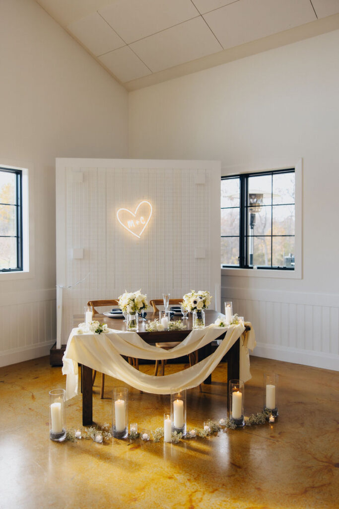 Intimate wedding dining setup with a heart-shaped neon sign above the table, surrounded by candles and white floral decorations