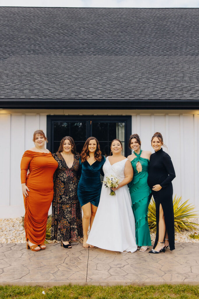 The bride in a classic white wedding dress stands with her bridal party, all wearing different colored dresses