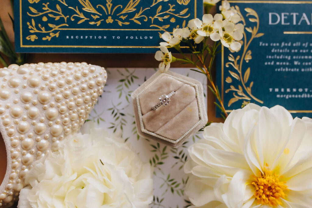 An engagement ring in a box next to wedding invitations, heels, and flowers. 