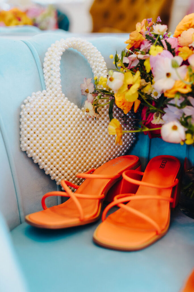 Orange heels on a blue couch next to a heart shaped purse and flowers. 