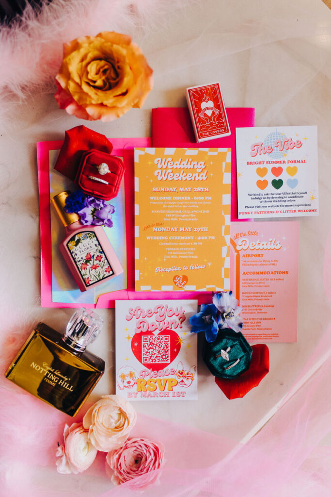 Brightly colored wedding invitations, jewelry, perfume, and flowers laid out together. 