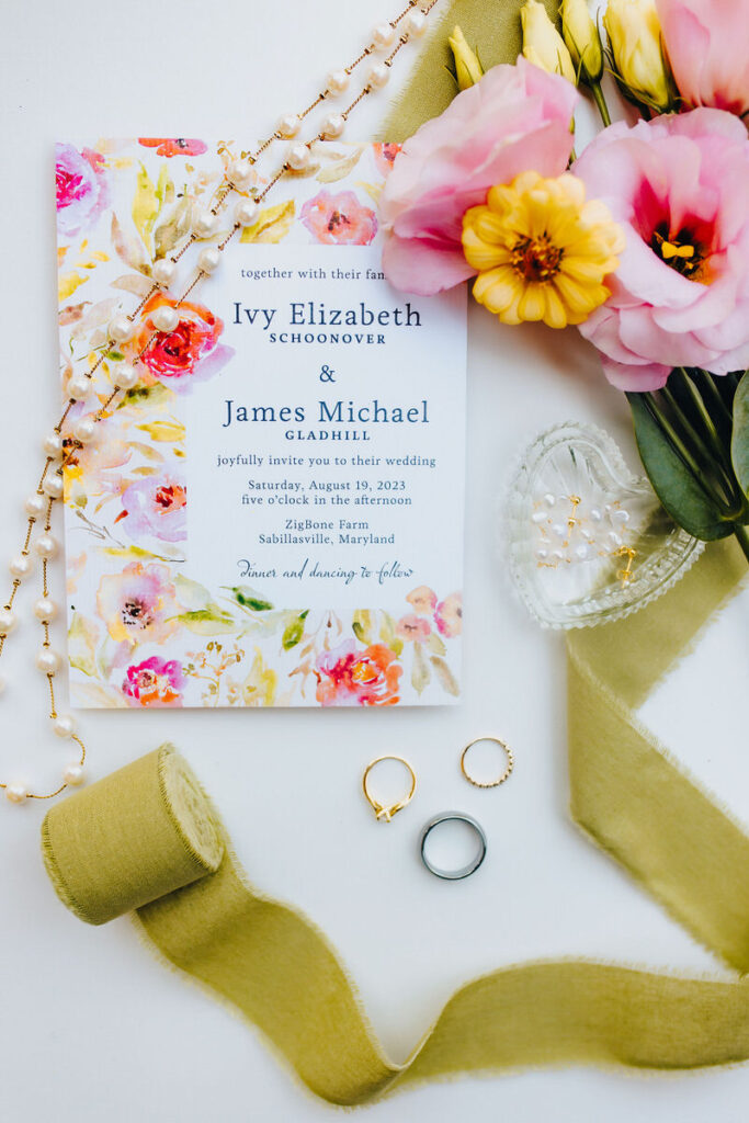 A wedding invitation and jewelry laid out next to flowers and ribbons. 