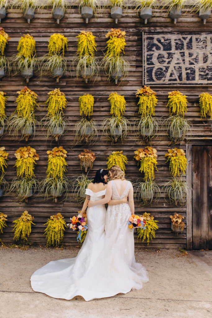 Two brides with their arms around each other in front of a plant wall
