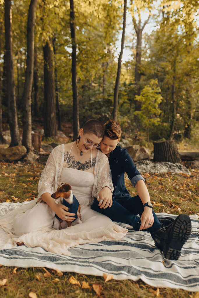 A couple sitting on a blanked in the woods looking down at a small dog in their lap. 
